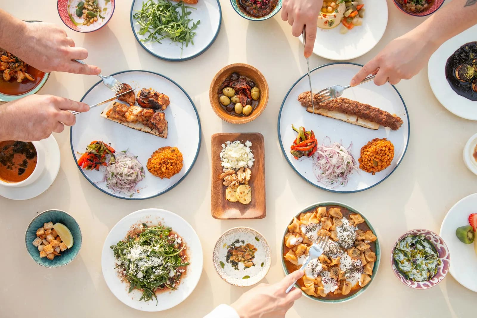 Yukarıdan çekilmiş ortak bir sofra: köfte ve şiş kebap tabakları, pilav, mezeler ve salatalar; birkaç el tabaklardan servis yapıyor.